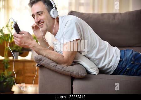 Happy man using a mobile listening to music lying on a sofa at home Stock Photo