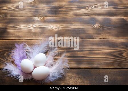 White chicken eggs on a wooden table in a nest of delicate pastel-colored feathers. Easter village background, natural eco-friendly products. Copy spa Stock Photo