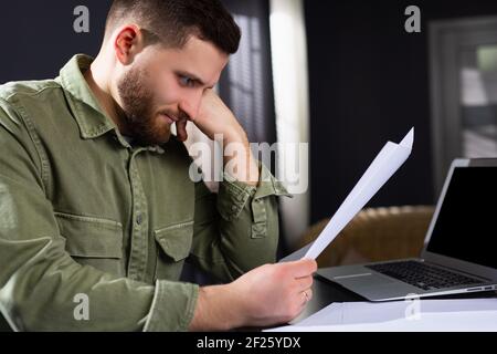 Caucasian man looking tired looking at the documents while sitting at the computer table. Documents, business and work online. Stock Photo