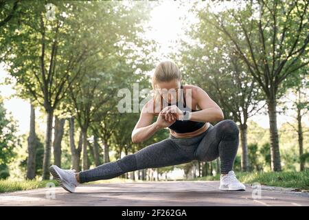 Endurance. Attractive middle aged athletic woman in sportswear looking focused, stretching her legs while training in a green park on a sunny day Stock Photo