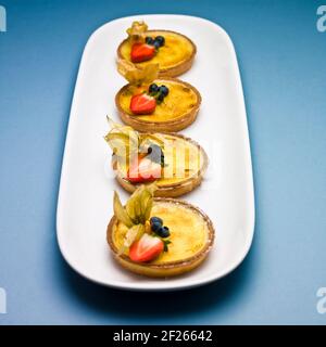 Four custard tarts, with blueberries, strawberries and physalis on a white dish on a blue background. Stock Photo