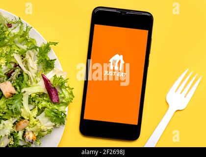 Just eat application icon on black screen of smartphone with lettuce salad plate and white plastic fork on yellow background Stock Photo