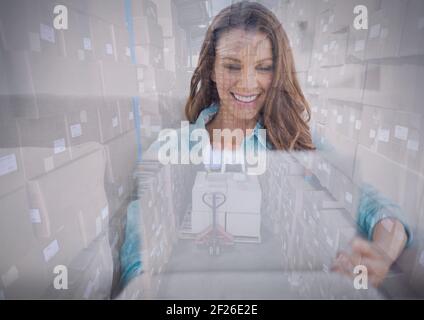 Composition of smiling woman using laptop over stack of cardboard boxes in warehouse Stock Photo