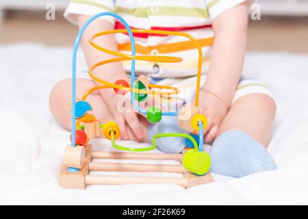 Unrecognizable caucasian baby playing with educational labyrinth. Sitting on white carpet in striped bodysuit.  Stock Photo