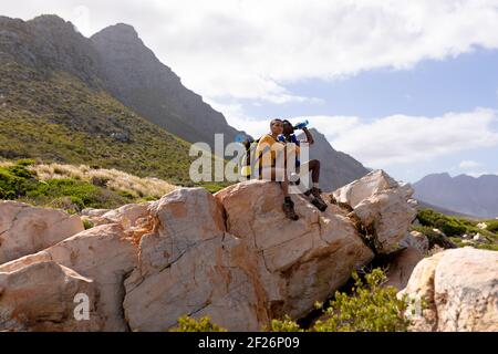 Fit afrcan american couple wearing backpack resting drinking water on the coast Stock Photo