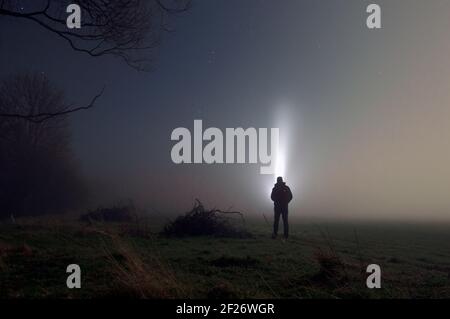 An atmospheric, moody concept. Of a man with a torch looking at the sky, in a field on a misty winters night. UK. Stock Photo