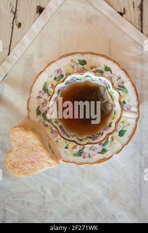 Cup of tea from above.  Close-up. Vintage tea cup and saucer with steeping teabag and heartshaped scone. On vintage white Irish linen. Stock Photo
