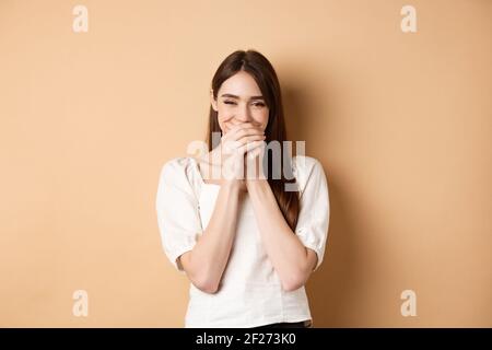 Happy girl laughing and covering mouth with hands, hiding her smile and chuckle over something funny, standing on beige backgrou Stock Photo