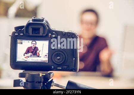 Video blogger at home: Camera on tripod is filming a young man sitting on his working place Stock Photo