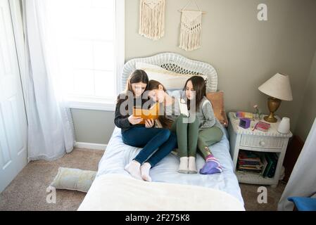 Three sisters reading on the bed together in a sunny bedroom. Stock Photo