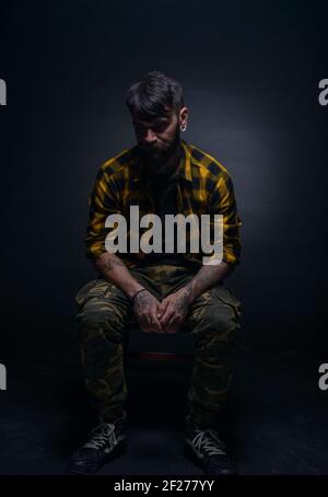 Handsome male model with tattoos and tartan shirt is sitting on a chair isolated on a black background Stock Photo