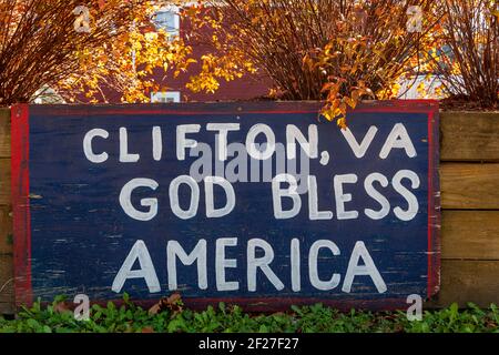 A blue painted wooden board attached to wooden planks near the train tracks has a notice on it that says Clifton, VA God Bless America. Clifton is a s Stock Photo