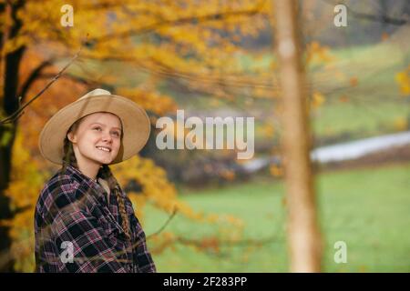 Young woman in a straw hat and plaid shirt smiling outdoors among autumn foliage. Stock Photo