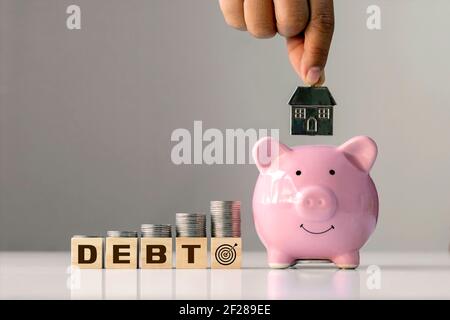 Concept of debt and financial goals.Coin on stacked wooden blocks labeled debt and hands holding house model in pink pig savings. Stock Photo