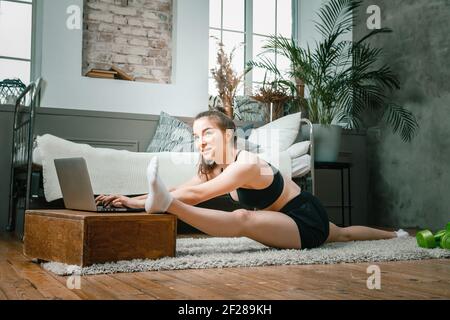 Positive athletic woman with neatly tied hair and tight sportswear stretching and doing  twine  at home. The young woman goes in for sports at home. Stock Photo