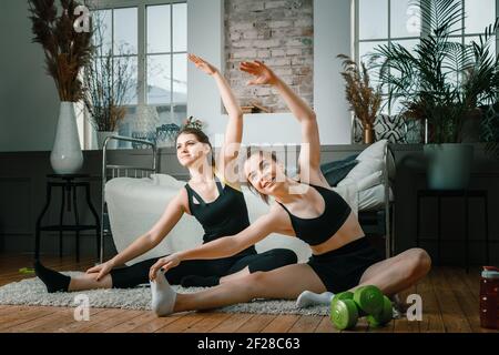 Positive athletic women with neatly tied hair and tight sportswear stretching at home. Girls go in for sports at home. Stock Photo