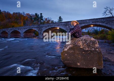 Young woman sitting on the rocks, downstream of the Pakenham Bridge in Ontario. a 5 span stone bridge that crosses the Mississippi river in Canada Stock Photo