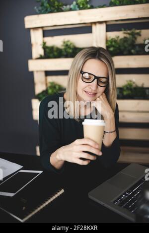 pretty, young and blond woman with stylish, modern black glasses sits in a sustainable office and enjoys a coffee to go Stock Photo
