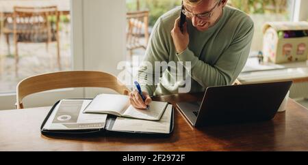 Man sitting at table talking on phone and writing in a book. Man working from  home with laptop and documents on table. Stock Photo