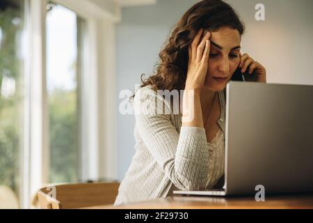 Woman looking stressed out while talking on a cellphone. Woman feeling stressed sitting at table with laptop and talking on mobile phone. Stock Photo