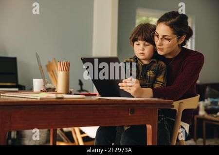 Woman and son sitting at table using digital tablet. Mother using digital tablet with son homeschooling. Stock Photo