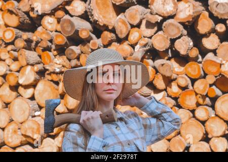 Outdoor portrait of young beautiful woman with long hair, ax in hand, wearing straw hat, in stylish lumberjack shirt on wooden background, people. Stock Photo