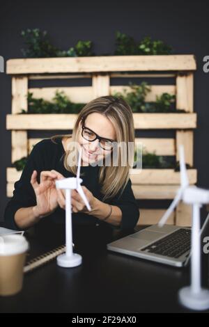 pretty, young woman with stylish, modern black glasses sits in a sustainable office and works with wind turbine models Stock Photo