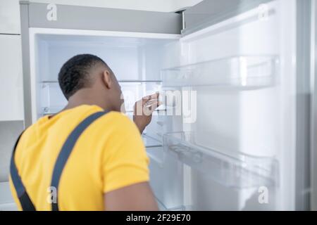 Dark-skinned service man in yellow tshirt repairing the fridge Stock Photo