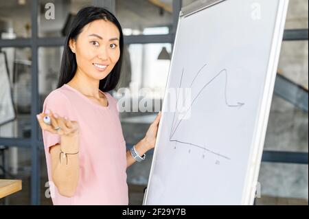 Modern pretty young Asian female school teacher streaming lesson online, professor, college tutor or coach looking at camera and holding a pen, drawing on the whiteboard and describing a graph Stock Photo