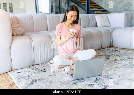 Smiling young Asian domestic woman with long black hair sitting on the floor and enjoying cup of tea coffee while watching movie, comedy show, series on laptop, spending leisure time alone at home Stock Photo