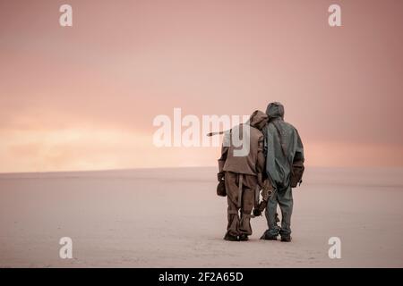 A man and a woman in chemical protection suits stand side by side on a sandy landscape. In the hands of gas masks. A post-apocalyptic world. Stock Photo
