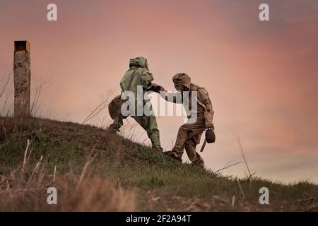 A man in a gas mask and a chemical protection suit helps another person to walk. Post-apocalypse after nuclear war and radiation pollution.  Stock Photo