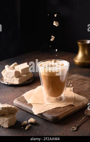 Tasty recipe coffee latte with halva. Sprinkles halawa in glass cup on dark brown background. Vertical. Close up. Stock Photo