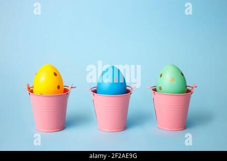 Three colored decorated eggs in buckets on a blue background. Easter greeting card in minimalist style concept.Copy space Stock Photo