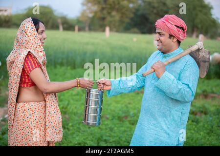 Happy traditional indian woman wearing sari giving tiffin box to his husband in agriculture field, happy farmer couple holding lunch box. Stock Photo