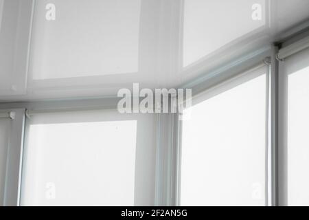 White fabric roller blinds on the plastic window on a balcony in the living room with a reflection in a stretch ceiling. Stock Photo