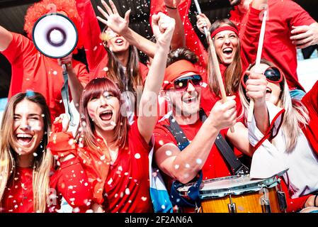 Football supporter fans cheering with confetti watching soccer match cup at stadium tribune - Young people group with red t-shirt having excited fun Stock Photo