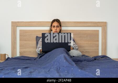 Young woman, in her thirties, teleworking with her laptop, sitting and covered by a blue duvet, in the comfort of her bedroom bed. Stock Photo