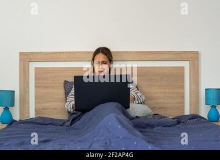 Young woman, in her thirties, teleworking with her laptop, sitting and covered by a blue duvet, in the comfort of her bedroom bed. Stock Photo