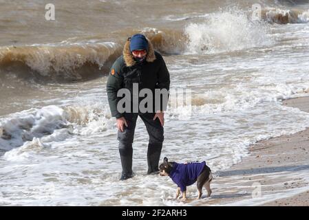 Southend on Sea, Essex, UK. 11th Mar, 2021. The strong overnight winds have continued into the morning, driving the mid-morning Thames Estuary high tide against the seawall. A few people braved the water. Male paddling in Wellington boots with dog Stock Photo