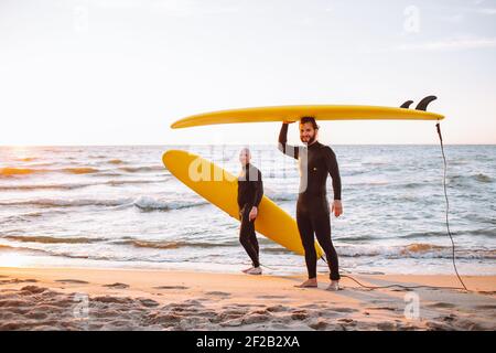 Two young surfers in black wetsuit with yellow surfing longboards at ocean coast at sunset. Water sport adventure camp and extreme swim on summer vaca Stock Photo