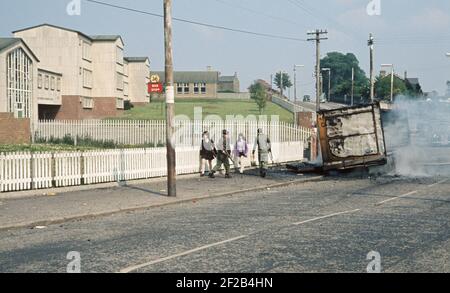 BELFAST, UNITED KINGDOM - JUNE 1972. School Children passing infront of British Army Soldiers and Burning Hijacked vehicle, Ballymurphy, West Belfast during The Troubles, Northern Ireland, 1970s Stock Photo