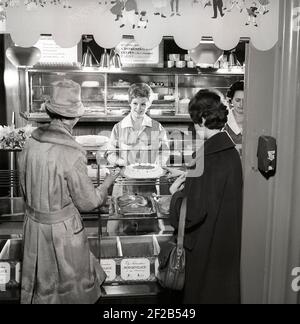 1960s coffee shop. Two women are standing in front of the counter that is full with freshly baked cakes and pastries. Sweden 1960s ref CX80-4 Stock Photo