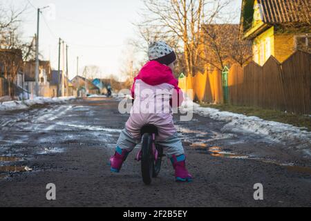 Child in pink jumpsuit rides balance bike on dirt road, in countryside Stock Photo
