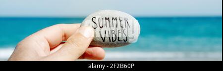 closeup of a caucasian man on the beach holding a stone, with the text summer vibes written in it, in front of the ocean, in a panoramic format to use Stock Photo