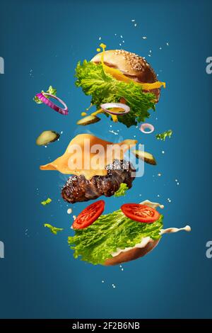 Hamburger with flying ingredients on blue studio background. Fast food concept. Bun, salad, meat, cheese and tomatos, onion in flight. Restaurant cuisine, advertising concept. Tasty, juicy, cheesy. Stock Photo