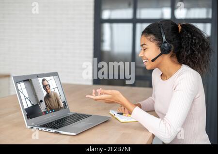 Video call. An African woman wearing headset and smart casual wear using laptop for talking with a male colleague, an online teacher or mate via video. Side view Stock Photo