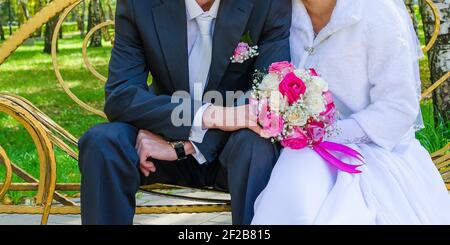 The groom and the snow-white bride are sitting on a yellow bench and holding a wedding bouquet of multi-colored roses in front of them. Stock Photo