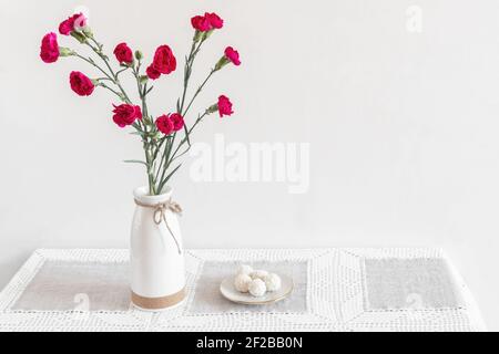 On the table in a ceramic vase are red carnations, next to a plate of sweets. White background, Scandinavian style. Front view, copy space. Stock Photo