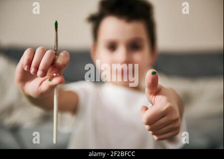 Image of a blurred schoolboy holding a paint brush in one hand and showing thumb up with other Stock Photo